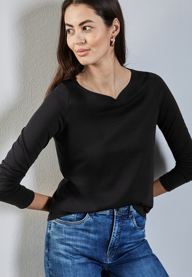 Schwarzes Langarm-Oberteil mit V-Ausschnitt, aus weichem Stoff gefertigt. Kombiniert mit blauen Jeans im klassischen Schnitt und mit leichter Waschung.