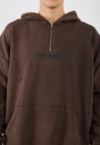 Ruda gobtuvė su priekiniais užtrauktukais, su juodu logotipu "URTRN". Pagaminta iš minkštos medžiagos, su laisva siluetu ir kengūros kišene.
