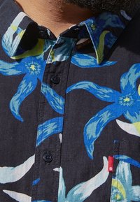 Chemise à manches courtes noire à motifs floraux bleus et verts, col boutonné et petite étiquette rouge sur la poche.
