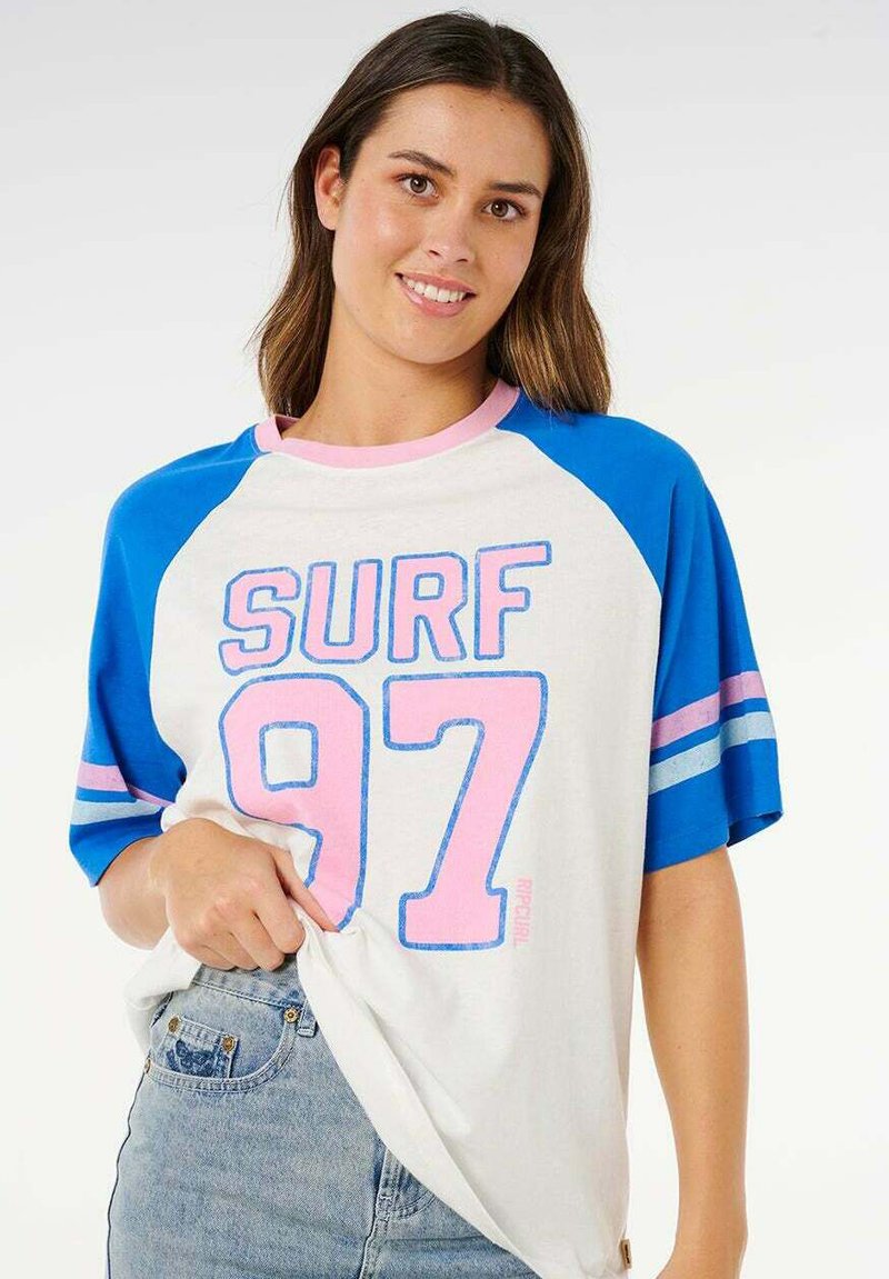 Jonge vrouw die lacht, gekleed in een wit T-shirt met blauwe mouwen en roze tekst "SURF 97," en lichtblauwe spijkerbroek.