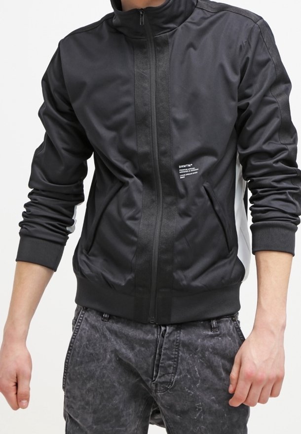 Personne portant une veste noire zippée avec des poignets côtelés et deux poches avant, assortie à un jean gris foncé délavé.