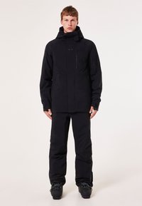 Veste noire imperméable avec un col haut, des poches zippées et une capuche ajustable, associée à un pantalon assorti et des bottes de ski.