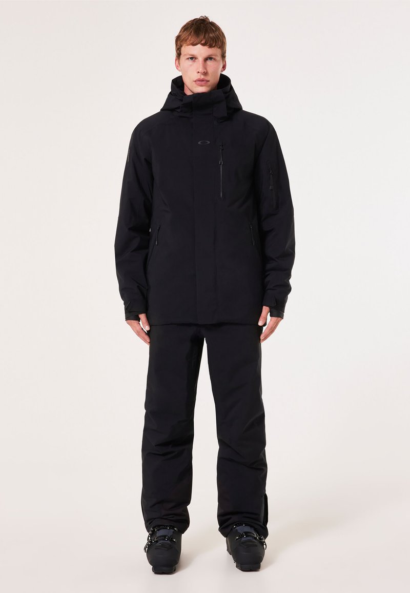 Schwarze wasserdichte Jacke mit hohem Kragen, Reißverschlusstaschen und verstellbarem Kapuzen, kombiniert mit passenden Hosen und Skischuhen.