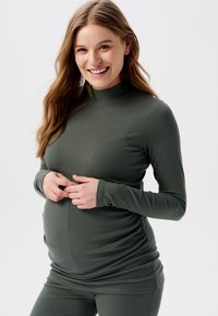 Mørkegrøn ribbet langærmet top med høj krave, der har samlet detalje i siderne, hvilket giver stræk og en tætsiddende silhuet.
