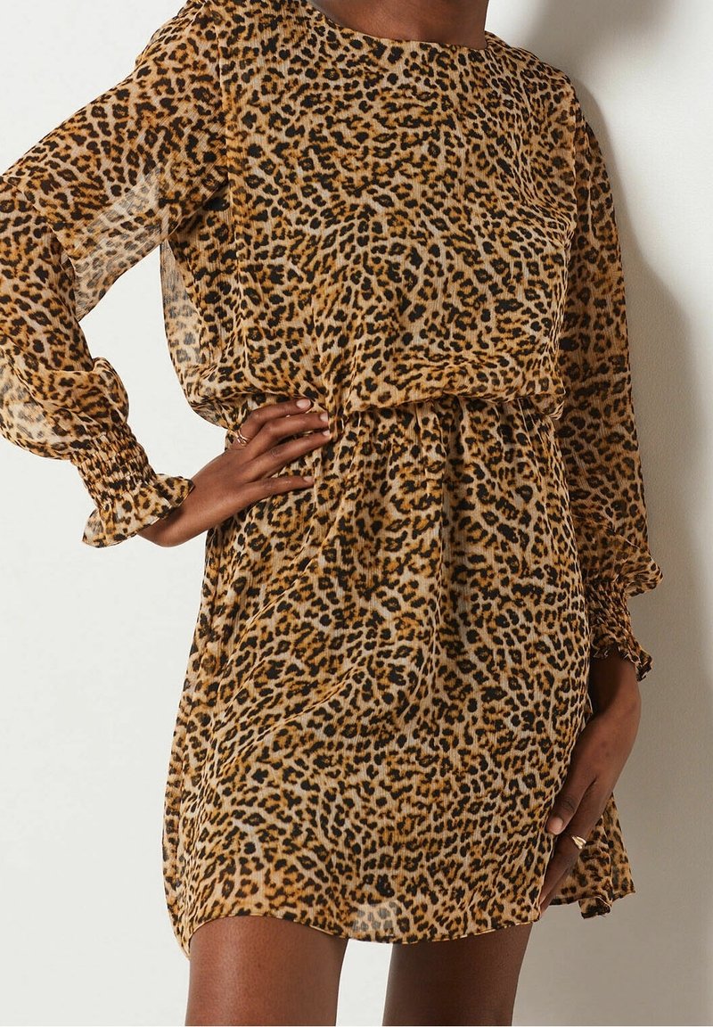 Femme portant une robe longue à manches longues en tissu transparent à imprimé léopard, taille élastique et poignets volantés, posant avec une main sur la hanche.