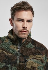 Sudadera de forro polar en patrón de camuflaje marrón, verde y negro; cuenta con un cuello alto, cierre de cremallera y un pequeño bolsillo en la parte delantera.