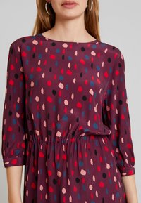 Fialové šaty s kulatým výstřihem a¾ rukávy, se staženým pasem. Mají vzor červených, modrých, černých a růžových teček.