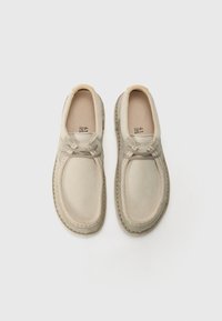 Beige slip-on sko laget av myk semsket skinn med rund tå, dekorativ søm og lett design. Utsmykket med lysegrå snører.