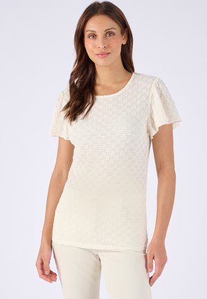 Vrouw met lang bruin haar die een kortemouwige, gestructureerde crèmekleurige blouse en lichte broek draagt tegen een effen achtergrond.