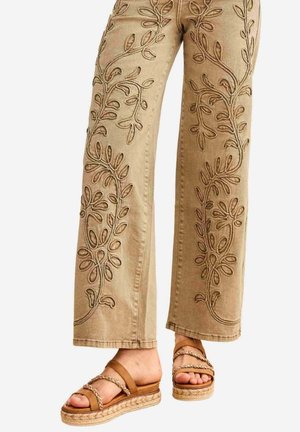Pantalones anchos beige con bordado floral en relieve, usados con sandalias de plataforma marrones con tiras trenzadas.