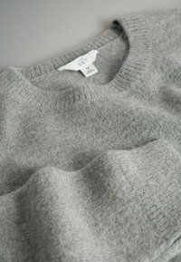 Szary sweter z dzianiny z okrągłym dekoltem, miękką teksturą oraz metką wskazującą rozmiar M. Tkanina ma delikatny połysk i subtelny wzór.