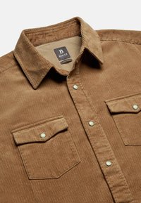 Bruine corduroy shirt met een button-down kraag, twee voorzakken met kleppen en parel drukknopen. Gestructureerde stof is ribbelig.