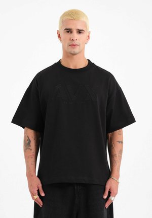 Joven con cabello corto decolorado, vistiendo una camiseta negra de gran tamaño con el logo AVX, mostrando múltiples tatuajes en los brazos y joyas, sobre un fondo blanco.
