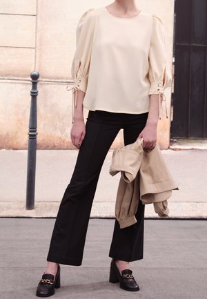 Personne portant un chemisier crème à manches bouffantes et un pantalon noir, tenant une veste beige, debout sur le trottoir près d'un mur en pierre et d'un poteau noir.