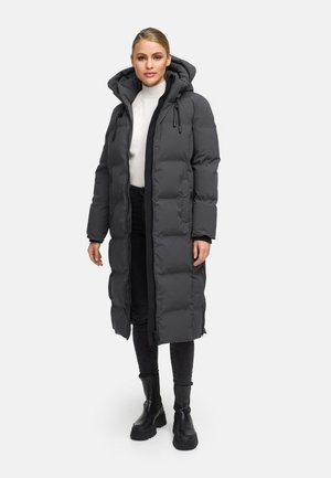 Manteau long et rembourré gris foncé, avec une capuche, une fermeture éclair à l'avant et des poignets élastiques. Associé à un pantalon noir et des bottes noires massives.