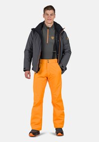 Giacca impermeabile grigia con dettagli arancioni, abbinata a pantaloni arancioni brillanti. Calzature scure completano l'outfit pronto per le attività all'aperto.