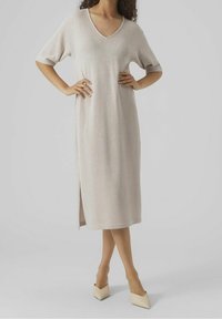 Knee-length beige knitted dress with a V-neck and short sleeves. Side slit detail, textured fabric, worn with pointed beige heels.