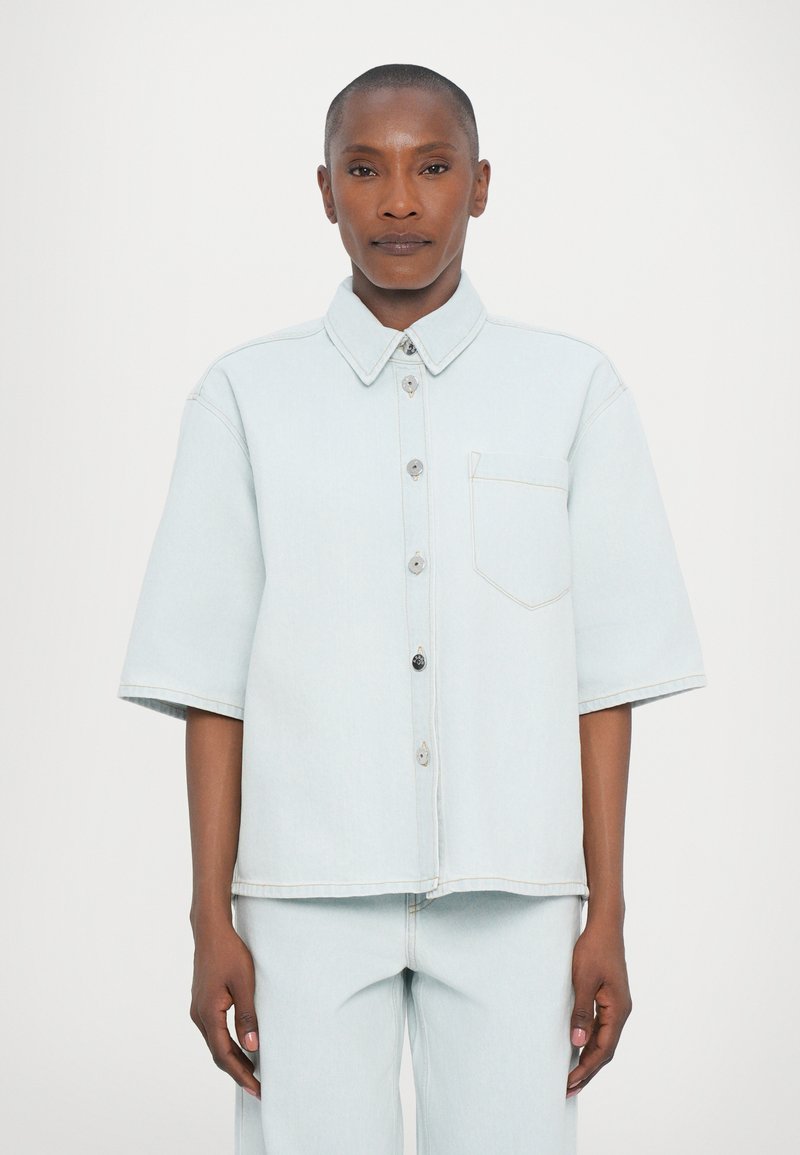 Camisa de ganga azul clara com mangas curtas, frente com botões, um único bolso no peito e costura em contraste. Textura suave, ajuste solto.