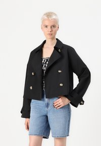 Giacca nera tagliata con design doppiopetto, bottoni argentati, colletto ampio e maniche arrotolate, abbinata a pantaloni shorts in denim e una maglietta a scacchi.