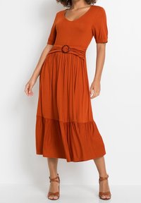 Rdzawopomarańczowa midi sukienka z miękkiego materiału z dopasowanym gorsetem, krótkimi bufiastymi rękawami i warstwową spódnicą, podkreślona okrągłym detalem paska.