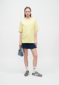 Mladá žena s tetováním na noze má na sobě žluté oversized tričko, námořnickou plisovanou sukni, bílé ponožky, šedé tenisky a drží modrou kabelku.