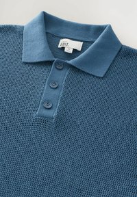 Blå strikket polo med en struktureret krave, tre-knaps lukning og tekstureret stof. Etiketten viser "NEXT EDIT" med størrelse "M."