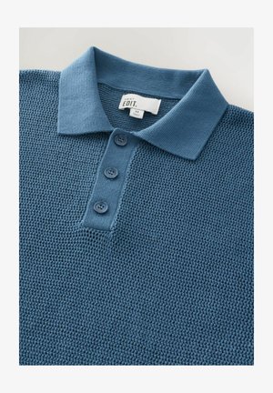 Modra pletena polo majica z strukturnim ovratnikom, tri gumbi in teksturirano tkanino. Oznaka prikazuje "NEXT EDIT" z velikostjo "M."
