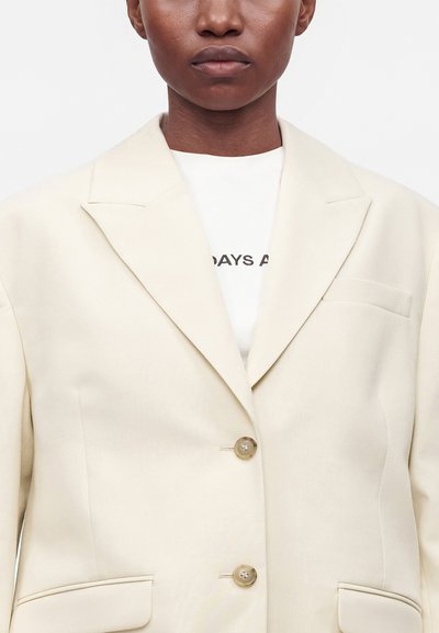 Personne portant un blazer beige clair à double boutonnage sur une chemise blanche avec un texte noir partiellement visible sur la poitrine.