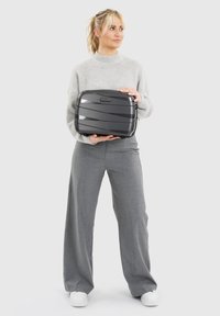 Vrouw in een grijs sweater en broek, met een kleine zwarte Redolz-koffer, die iets opzij kijkt, tegen een eenvoudige witte achtergrond.