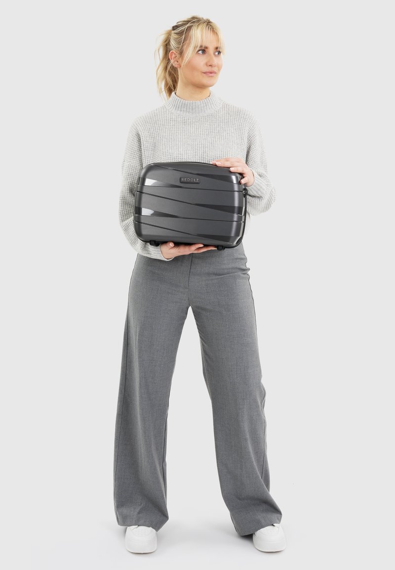 Vrouw in een grijs sweater en broek, met een kleine zwarte Redolz-koffer, die iets opzij kijkt, tegen een eenvoudige witte achtergrond.