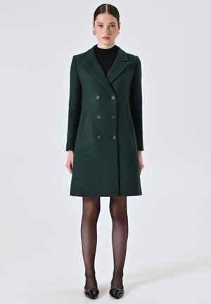 Grün melierter, doppelter Mantel mit Reverskragen, sechs Knöpfen und seitlichen Taschen. Stylisiert mit schwarzen Strumpfhosen und glänzenden schwarzen Schuhen.