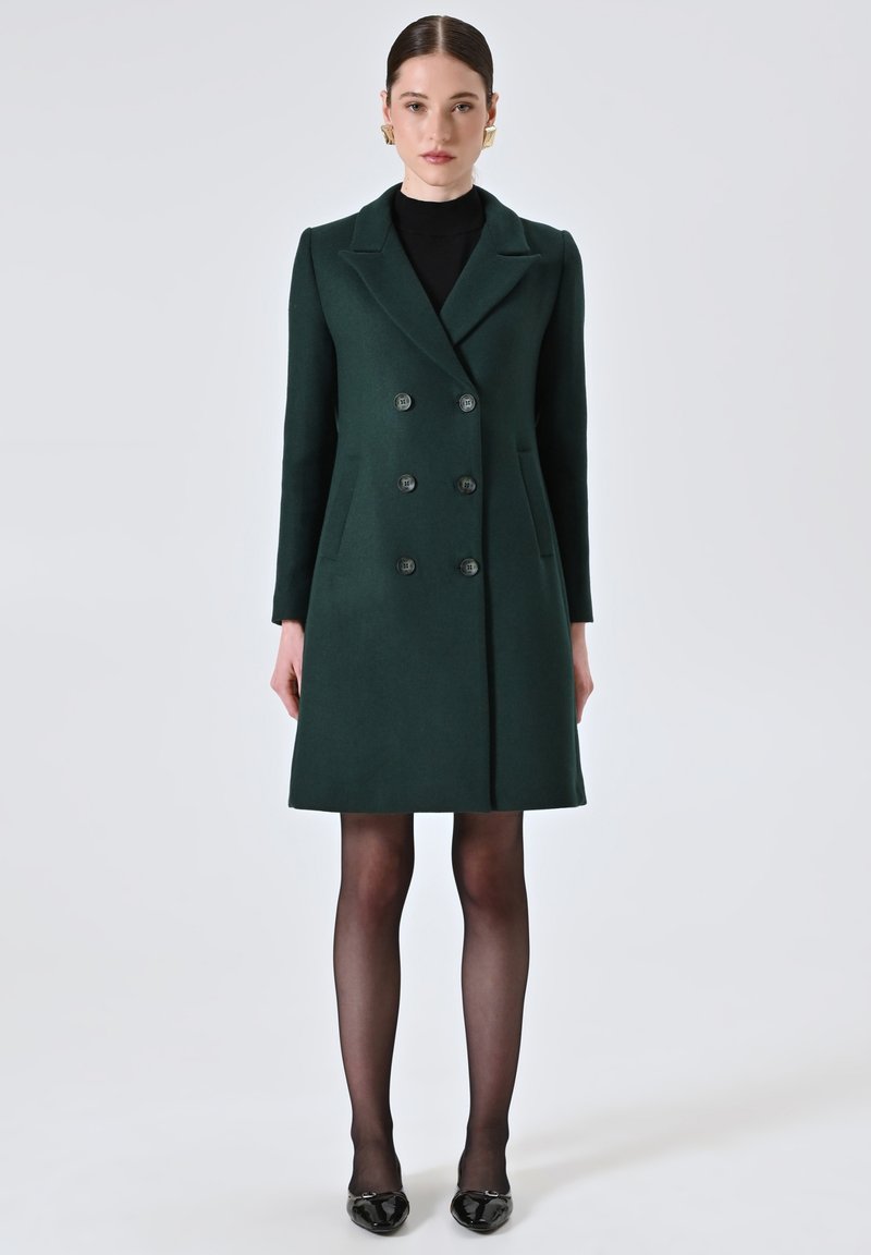 Cappotto doppiopetto in misto lana verde, con rever a punta, sei bottoni e tasche laterali. Abbinato a collant neri e scarpe nere lucide.