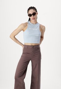 Top court en bleu clair avec texture côtelée et logo brodé blanc, associé à un pantalon marron taille haute et large. Lunettes de soleil noires.