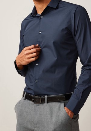 Homme portant une chemise bleu foncé boutonnée et un pantalon gris avec une ceinture noire, une main ajuste la chemise et l’autre dans la poche.