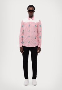 Chemise rose à manches longues avec broderie florale, empiècement et col blanc ; associée à un pantalon skinny noir et des baskets blanches. Le modèle porte des lunettes de soleil.