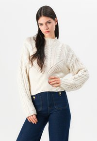 Krém színű, kábelmintás pulóver magas nyakkal, bordás mandzsettákkal és rövidített alsó résszel, sötétkék, magas derekú farmernadrággal párosítva.