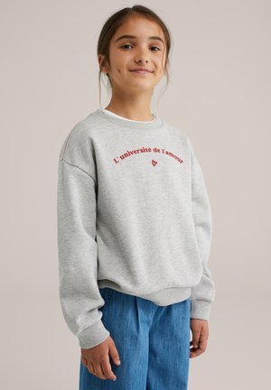 Grauer Sweatshirt mit rotem Text „L’université de l’amour“ und Herzdetail. Mit gerippten Bündchen und Saum ausgestattet, kombiniert mit blauen, entspannten Hosen.