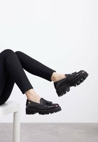Zapatos de charol negros con suela gruesa texturizada y detalle decorativo de borlas. Posicionados sobre piernas que usan legging negros.