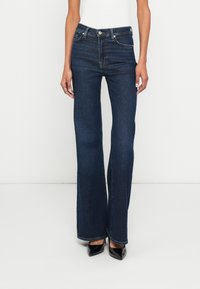 Jean évasé bleu foncé à taille haute, avec cinq poches et des surpiqûres contrastantes. Confectionné en denim avec une texture douce et un design minimaliste.