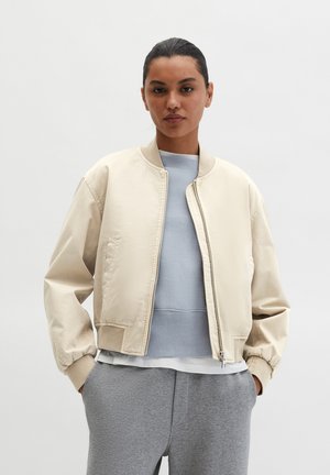 Beige Bomberjacke mit Reißverschluss, gerippten Bündchen und lockerem Schnitt, getragen über einem hellblauen Sweatshirt und grauen Jogginghosen.