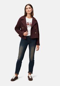 Burgunderfarbenes, tailliertes Doppelreiher-Jackett, weißes Grafik-T-Shirt, dunkelblaue Jeans, braune Sneakers mit weißen Schnürsenkeln, stehende Pose, einfarbiger Hintergrund.