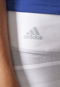 Primer plano del torso de una persona que lleva una camiseta azul y pantalones cortos Adidas blancos con rayas grises punteadas y el logo de Adidas en la parte frontal.