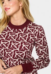 Pull en maroon et crème à motifs avec un col rond côtelé et des poignets côtelés. Le tissu présente un design complexe de formes superposées.