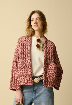 Woman with shoulder-length hair wearing patterned pink quilted jacket, white graphic t-shirt, blue jeans, woven belt, and sunglasses clipped to shirt.