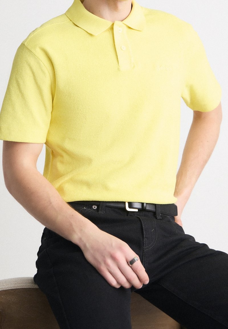 Homme assis portant un polo à manches courtes jaune vif, un pantalon noir avec une ceinture noire, et une bague en argent à la main droite.