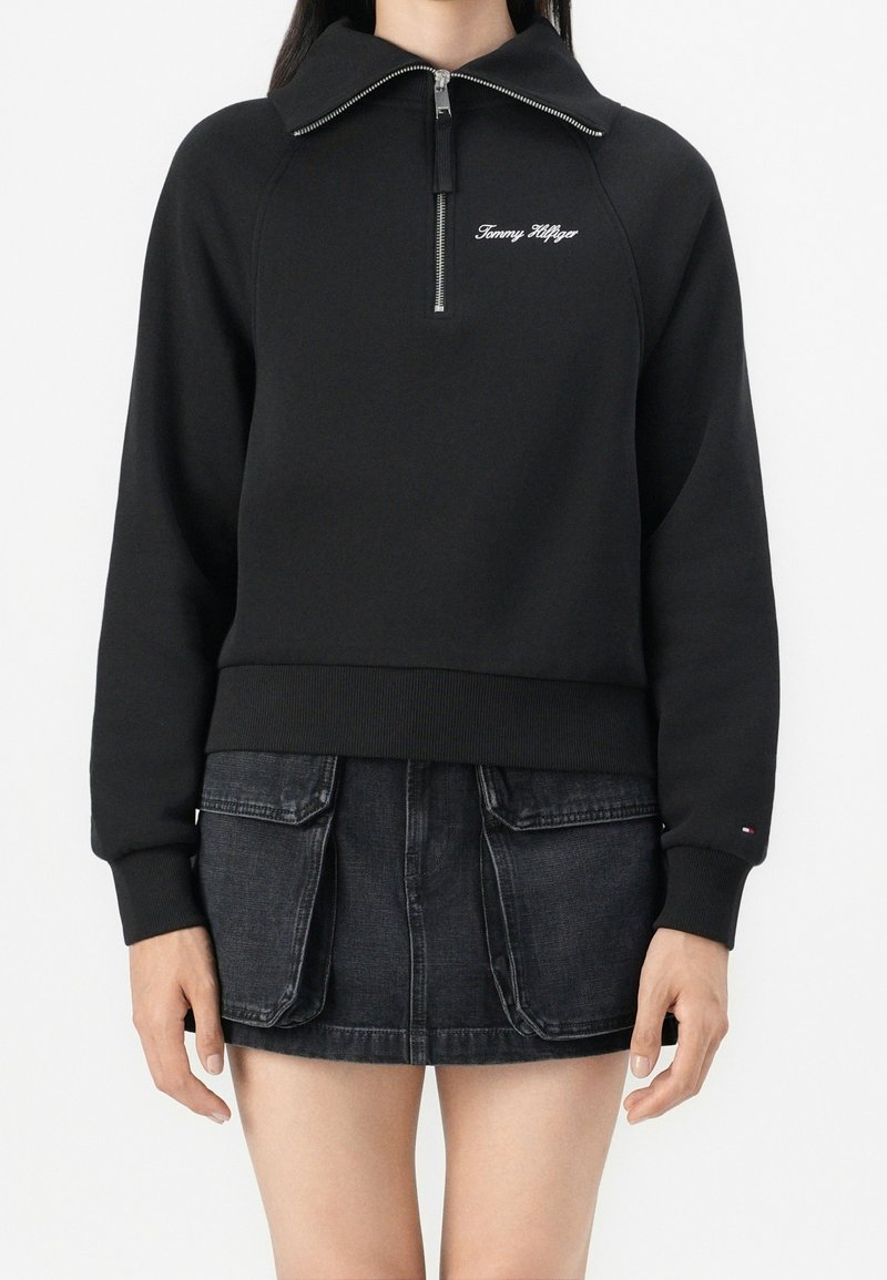 Personne portant un sweat-shirt noir à demi-fermeture éclair avec le logo "Tommy Hilfiger" et une mini-jupe en denim foncé avec de grandes poches avant.