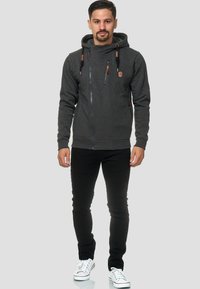 Sudadera con cremallera en gris oscuro y cuello alto, detalles de cuero y cordones ajustables, combinada con jeans negros y zapatillas blancas.