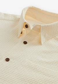 Chemise beige clair à boutons avec un tissu texturé, col écarté et boutons marron. Comprend une poche unique sur la poitrine.