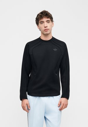 Ung mand iført en sort Adidas sweatshirt med lange ærmer og lyseblå bukser, stående mod en ensfarvet hvid baggrund.