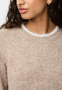 Ženska s kodrasto rjavo lasmi, oblečena v bež puhasto pleteno pulover z belim robom na vratu in srebrnim obročkom v ušesu.